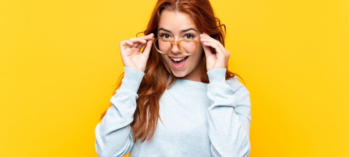A young woman excited while pulling her glasses off her face with a yellow background