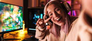 A young woman giving a peace sign and smiliing while taking a photo at her gaming desk in her room.