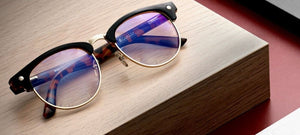 a pair of clubmaster gaming glasses sitting on a tan block of wood with a ran background
