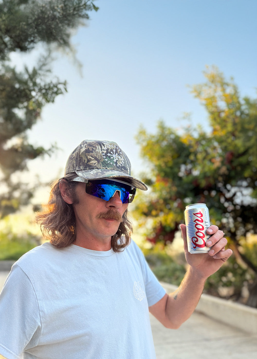 Man holding a Coors light beer can outdoors with trees and clear sky in the background